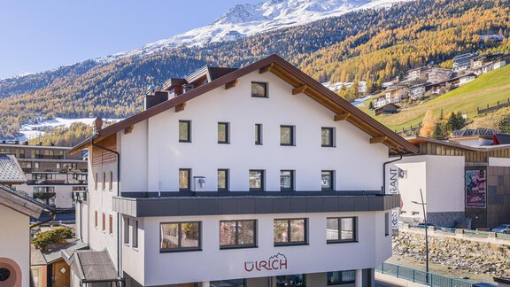 A modern building with the inscription "URICH" in the foreground. In the background, snow-covered mountains and autumn trees can be seen. - Haus Ulrich - Sölden