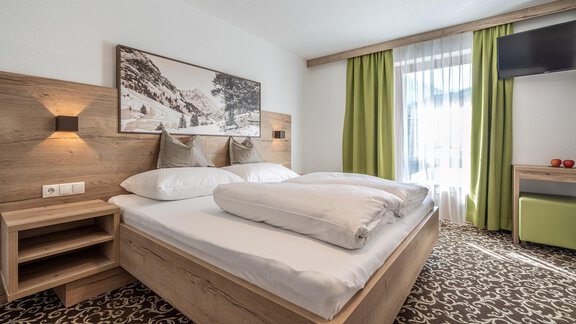 A modern bedroom with a large bed and a wooden-paneled headboard. The curtains are green, and there is a nightstand next to the bed. - Haus Ulrich - Sölden