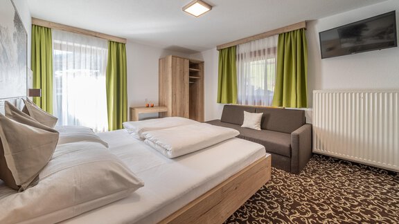 A modern room with a comfortable double bed and a sofa bed. Large windows with green curtains allow for plenty of light. - Haus Ulrich - Sölden
