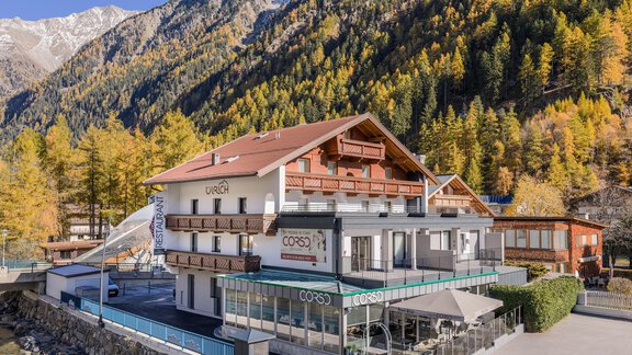 A modern hotel by the riverbank, surrounded by majestic mountains. The trees are colored in autumn foliage, giving the scene a picturesque atmosphere. - Haus Ulrich - Sölden