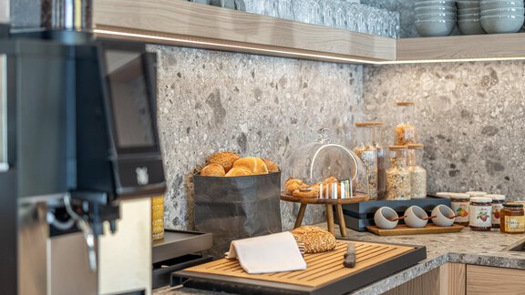 A modern kitchen with a stone countertop and elegant wooden shelves. Buns, muesli, and various jams are displayed on the counter. - Haus Ulrich - Sölden