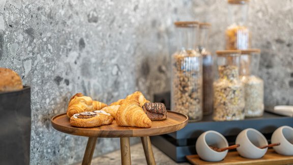 A wooden tray with fresh croissants and a small sweet treat is on a gray countertop. In the background, glass containers with various cereals can be seen. - Haus Ulrich - Sölden