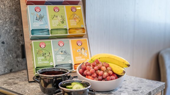 A table with various tea varieties in colorful packaging and a bowl of fresh fruit, including grapes and bananas. Next to it are two small bowls with additional snacks. - Haus Ulrich - Sölden