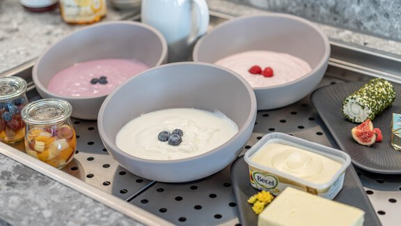 Three bowls of yogurt in different flavors are on a tray. Alongside, there is fresh fruit in glasses and an assortment of butter and cheese. - Haus Ulrich - Sölden