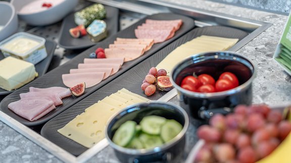 A selection of cold cuts, cheese, and fresh vegetables on a table. There are also grapes and cherry tomatoes in small bowls. - Haus Ulrich - Sölden