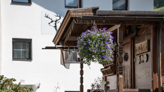 A traditional wooden house with a blooming flower pot. In the background, a modern building featuring a deer antler motif can be seen. - Haus Valtelas - Sölden