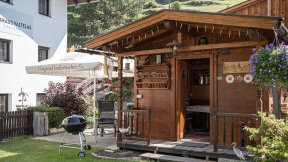A cozy wooden shed with an outdoor grilling station. In the background, there are green hills and a blue sky. - Haus Valtelas - Sölden