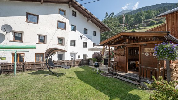 A cozy garden with a wooden house and a swing bench. In the background, a traditional building and the surrounding mountains can be seen. - Haus Valtelas - Sölden