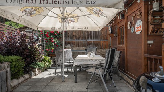 A cozy terrace with a large sun umbrella that advertises Italian ice cream. There is a table and chairs as well as blooming plants in the background. - Haus Valtelas - Sölden
