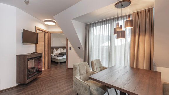 A modern living room with a dining table, two chairs, and a television on the wall. Large windows let in plenty of daylight and offer a view of a cozy sleeping area. - Home4You - Sölden