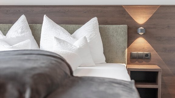 A cozy bedroom with a large bed, decorated with white pillows and a gray blanket. On the wall, there are warm light sources and a nightstand with power outlets. - Home4You - Sölden