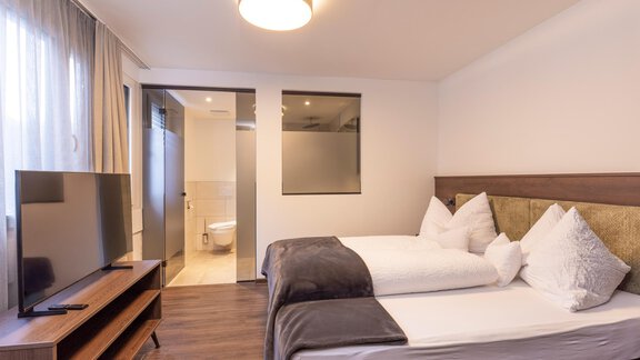 A modern apartment room with two beds and a flat-screen TV. In the background, there is a bathroom with a frosted glass wall. - Home4You - Sölden