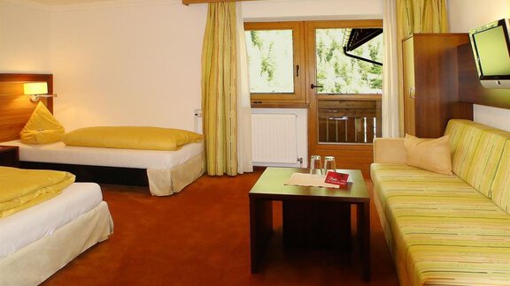 A cozy hotel room with two beds and a couch. Large windows provide a view and daylight. - Hotel Garni Hainbacherhof - Sölden