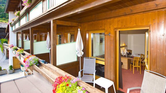 A cozy balcony with flowers and chairs, overlooking a wooden building. In the background, a bright room with a table and chairs can be seen. - Hotel Garni Hainbacherhof - Sölden