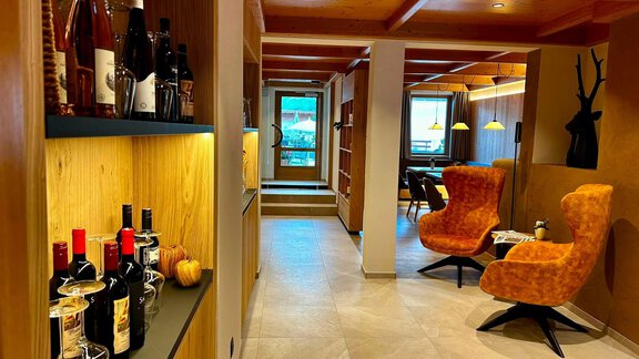 A modernly designed reception area with wooden shelves and cozy orange armchairs. On a shelf, several bottles of wine and a short table are decorated. - Hotel Garni Hainbacherhof - Sölden