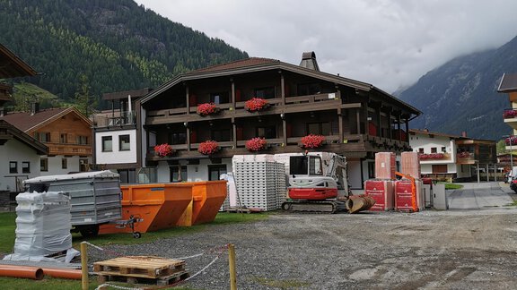 A construction site in a alpine region with a multi-story building in the background. There are building materials and machinery on the site, surrounded by mountains and clouds. - Hotel Garni Maria Theresia - Sölden
