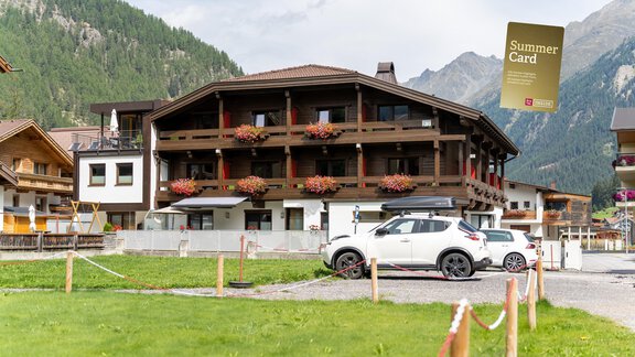 A beautiful holiday home in the Alps with many flowers on the balcony. In the foreground, there is a car in a parking lot. - Hotel Garni Maria Theresia - Sölden