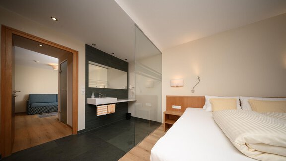 A modern room with a double bed and a bathroom with a glass wall. In the background, another room with a sofa can be seen. - Hotel Garni Maria Theresia - Sölden