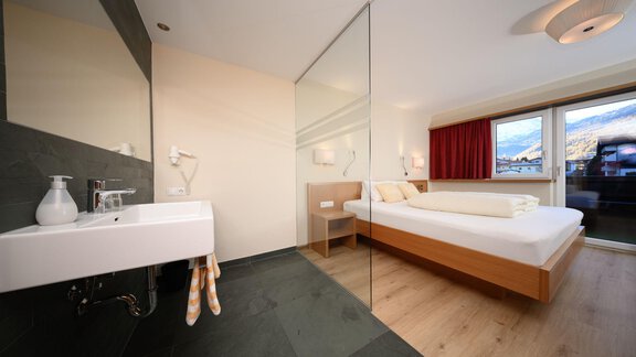 A modern bedroom with a large window and a view of the mountains. The bathroom is separated from the room by a glass wall. - Hotel Garni Maria Theresia - Sölden