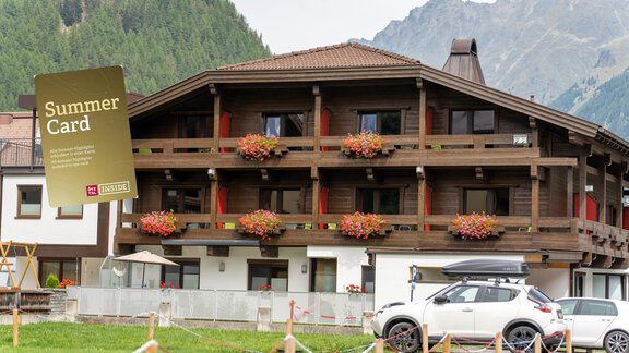 A cozy hotel with wooden balconies and blooming plants. In the foreground, there is a car, and in the background, mountains can be seen. - Hotel Garni Maria Theresia - Sölden