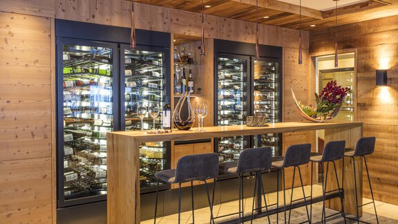 An elegant wine bar with wooden paneling and several cooling shelves. The bar is equipped with modern chairs and decorated with plants. - Hotel Hubertus - Sölden