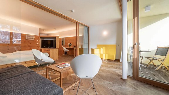 A modern room with wooden paneling and large windows. There is a sofa, a table, and access to a balcony. - Hotel Liebe Sonne - Sölden