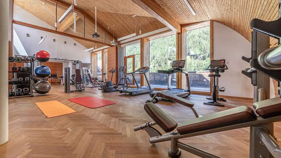 A modern gym with a wooden ceiling and large windows. There are various training equipment and mats on the floor. - Hotel Liebe Sonne - Sölden