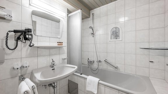 A modern bathroom with white tiles, a bathtub, and a shower. There is a sink with a mirror and towels. - Hotel Sölderhof - Sölden