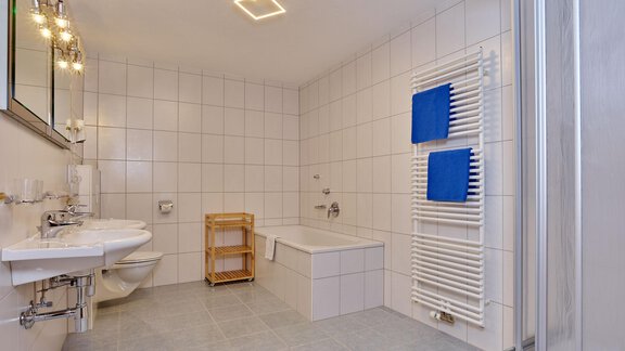 Ein modernes Badezimmer mit weißen Fliesen und hellen Lichtern. Es gibt eine Badewanne, zwei Waschbecken und Handtücher in blau. - La Luna - Sölden