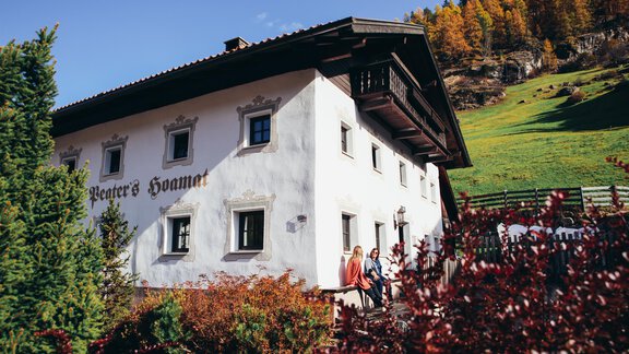 A picturesque house in nature with green meadows and colorful autumn foliage. Two people are standing in the foreground and enjoying the view. - Landhaus Peater's Hoamat - Sölden
