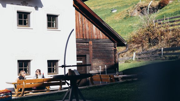 A rustic house made of wood with a green meadow in the background. Two people are sitting at a table outdoors, enjoying the surroundings. - Landhaus Peater's Hoamat - Sölden