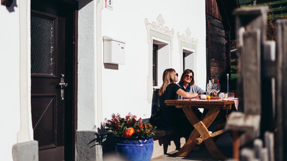 Two women are sitting at a wooden table in front of a house. They are enjoying a relaxing time outdoors with flowers and sunlight in the background. - Landhaus Peater's Hoamat - Sölden
