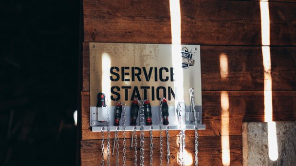 A sign with the inscription "SERVICE STATION" hangs on a wooden wall. Below are chains and hooks attached. - Landhaus Peater's Hoamat - Sölden