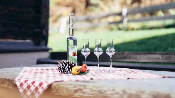 A decorated table with a bottle of schnapps and three glasses. The table is adorned with a checked cloth and a small selection of fruit. - Landhaus Peater's Hoamat - Sölden