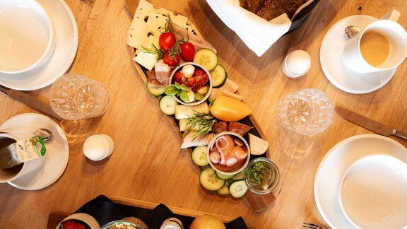 A beautifully arranged breakfast buffet with fresh fruits, cheese, and cold cuts on a wooden board. Around the buffet, there are empty plates and glasses on a wooden table. - LENI MOUNTAIN CHALET - Sölden