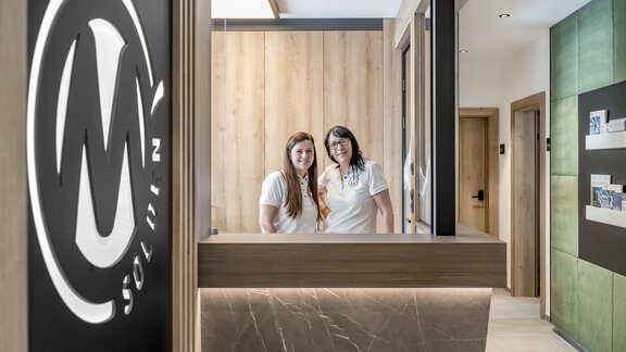 Two smiling women are standing at the reception desk of a modern hotel. In the background, you can see elegant wood and green tones. - Marco's Mountain Home - Sölden