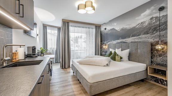 A modern room with a cozy bed and a small kitchenette. The wall is decorated with an impressive mountain motif and large windows provide plenty of daylight. - Marco's Mountain Home - Sölden