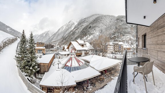 A winter landscape with snow-covered mountains and a charming village. On a balcony, there is a table and chairs with a view of the snowy surroundings. - Marco's Mountain Home - Sölden