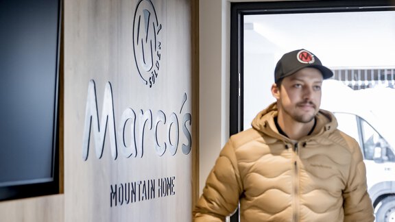 A man stands in front of a wall with the inscription "Marco's Mountain Home". The interior decor conveys a cozy atmosphere. - Sölden