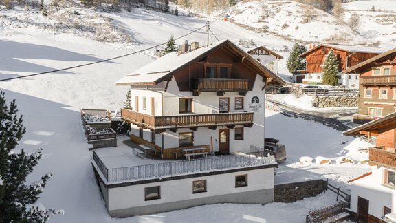 A large, white house in the snow, surrounded by snow-covered hills. In the background, more traditional buildings and trees are visible. - Mountain Lodge Sölden - Sölden