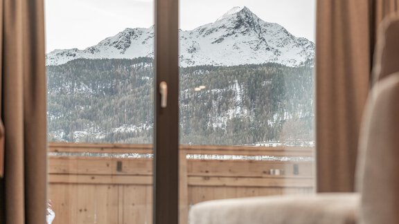 A view of snow-covered mountains through a glass door. In the foreground, there is a wooden railing and a cozy seating area. - Pension Miramonte - Sölden