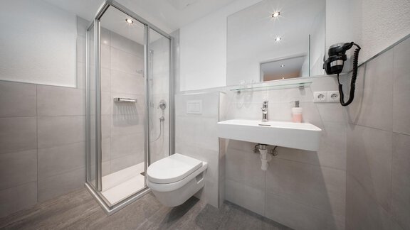 A modern bathroom with a shower, a sink, and a toilet. The walls are tiled in light gray and there is a large mirror. - Pension Pitze - Sölden