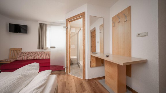 A modern room with wooden flooring, a bed, and a red sofa. Adjacent is a bathroom and a large mirror. - Pension Pitze - Sölden