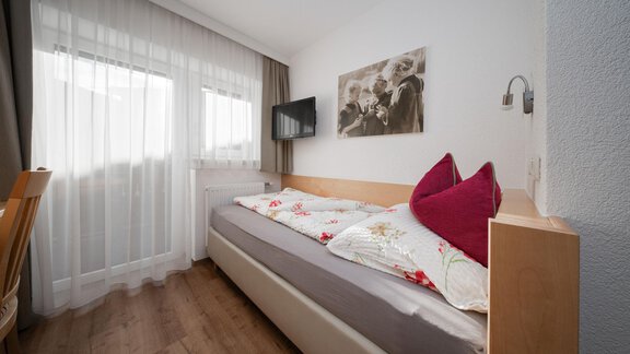 A cozy bedroom with a single bed, a television, and a large window. The walls are light, and on the bed are decorative cushions. - Pension Pitze - Sölden