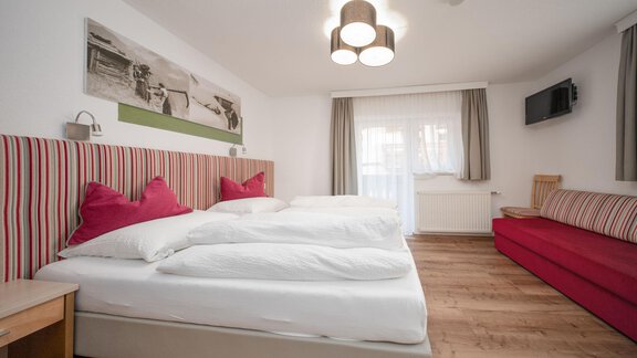 A modern bedroom with two beds, red cushions, and a cozy sofa. Bright walls and large windows create a friendly atmosphere. - Pension Pitze - Sölden