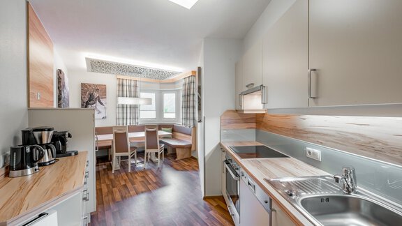 A modern kitchen with wooden surfaces and elegant appliances. In the background, there is a dining table with chairs and large windows. - Residenz Allegra - Sölden