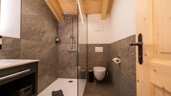 A modern bathroom with a glass shower and wood details. The walls are in neutral gray tones and there is a practical shelf space. - Selda Chalet - Sölden