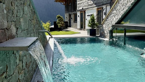 A modern swimming pool with clear water and a waterfall. In the background, there is a wooden house and lots of greenery. - The Peak Sölden - Sölden