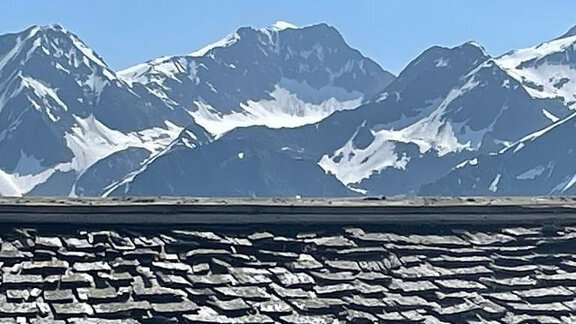 A mountain landscape with snow-covered peaks under a clear blue sky. In the foreground, a roof made of slate shingles is visible. - Top Apart Daniela - Sölden