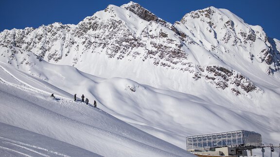 A snowy mountain landscape with tall peaks and a clear blue sky. In the foreground, skiers and a modern chalet can be seen. - Top Apart Daniela - Sölden
